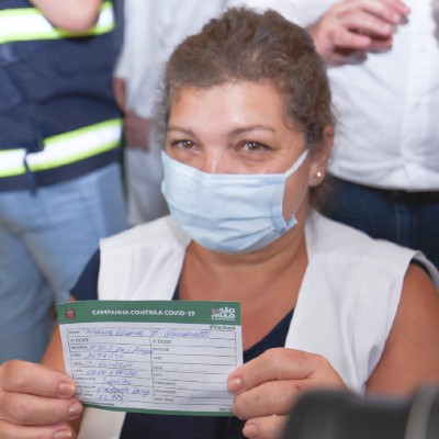 Foto de Maria Eliana Bonviccini Quiaratti. Ela está de máscara e com uma expressão de alegria. Segura com as duas mãos o comprovante da vacinação contra a Covid-19